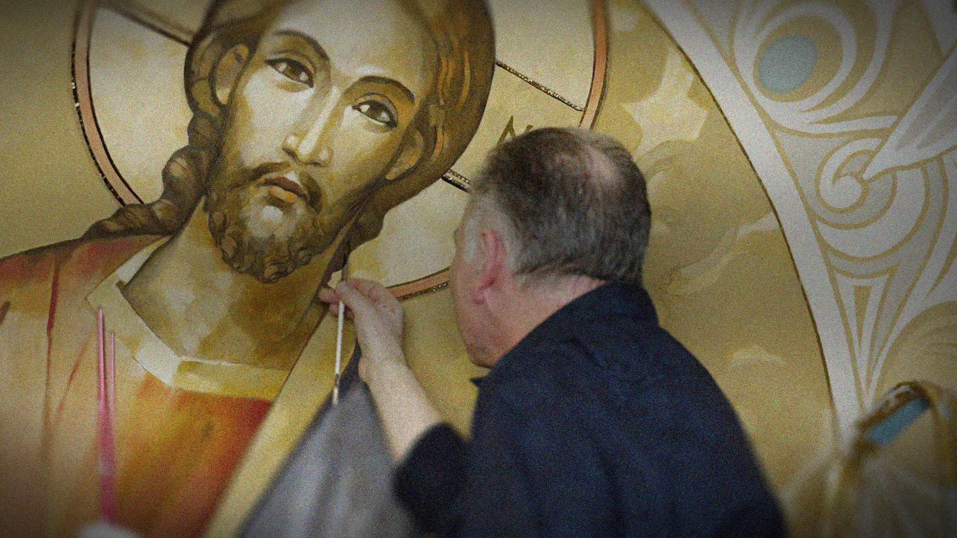 Das Bild zeigt Künstler Christof Häfliger, wie er in der Kirche St. Karl Borromäus der Priesterbruderschaft St. Pius X. ein Bild von Jesus Christus als Allherscher (Pantokrator) malt. Über dem Bild ist der Video-Titel «Behind The Scenes, Kirchenmalerei in der Kirche St. Karl Borromäus (FSSPX)» zu lesen.