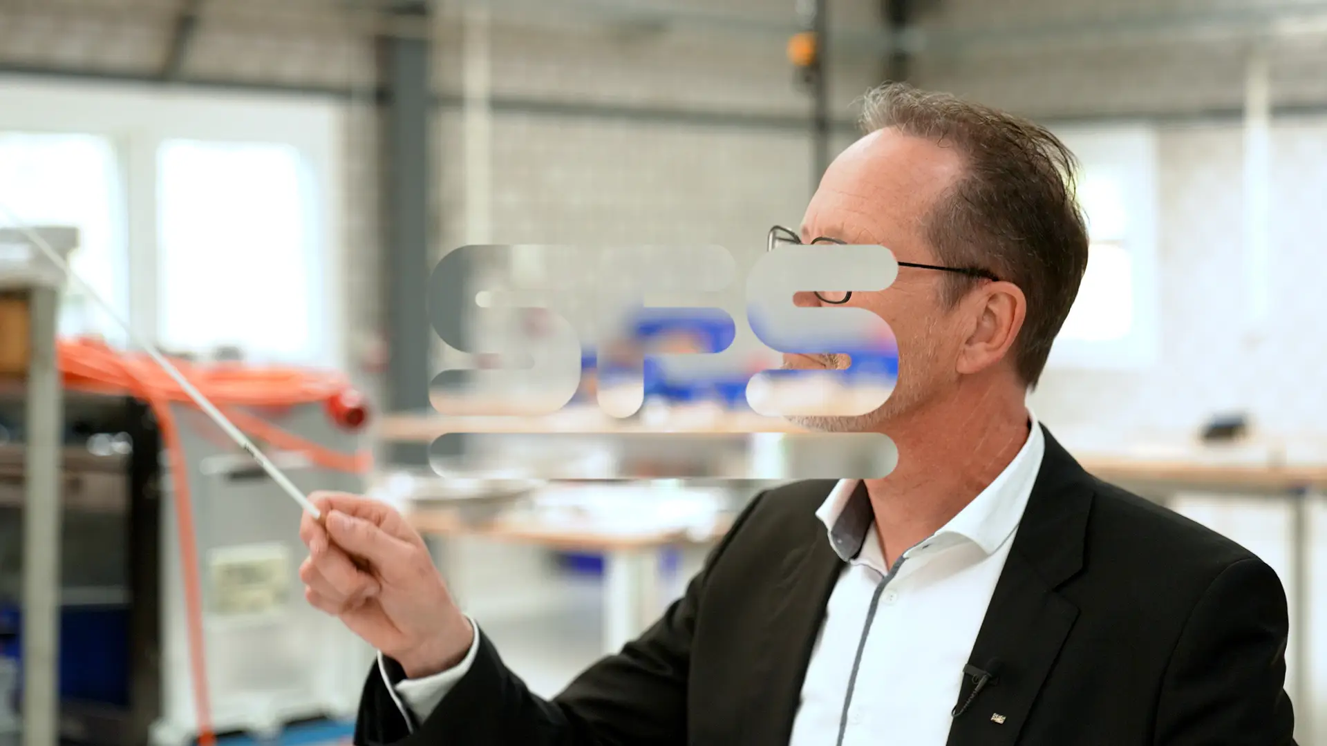 Ein Mann im Anzug dirigiert etwas in einer Lagerhalle. Im Vordergrund prangt der Schriftzug «SFS» (das Logo der Firma SFS).